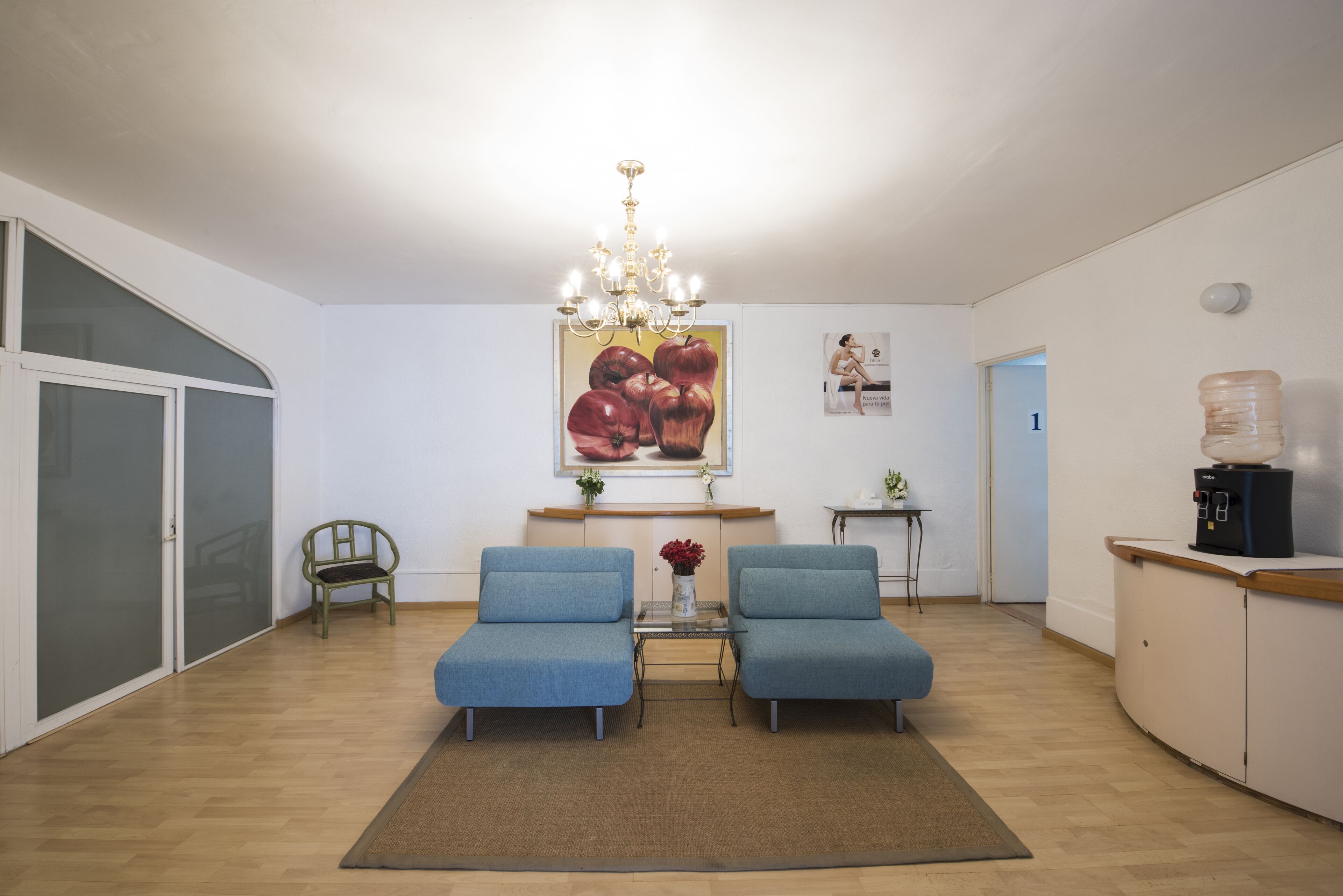 A spacious living area with two blue armchairs, a glass coffee table, a chandelier, and a large fruit painting on the wall.