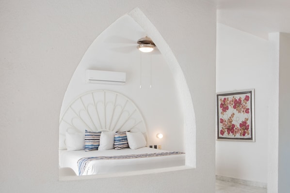 Un dormitorio con un techo abovedado único, una cama con almohadas a rayas, un aire acondicionado de pared y una obra de arte floral enmarcada.