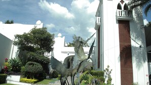 Une fontaine ornée d’une statue de dragon, située dans un bassin circulaire, entourée d’une végétation luxuriante et avec, en arrière-plan, un bâtiment blanc surmonté d’une tour.