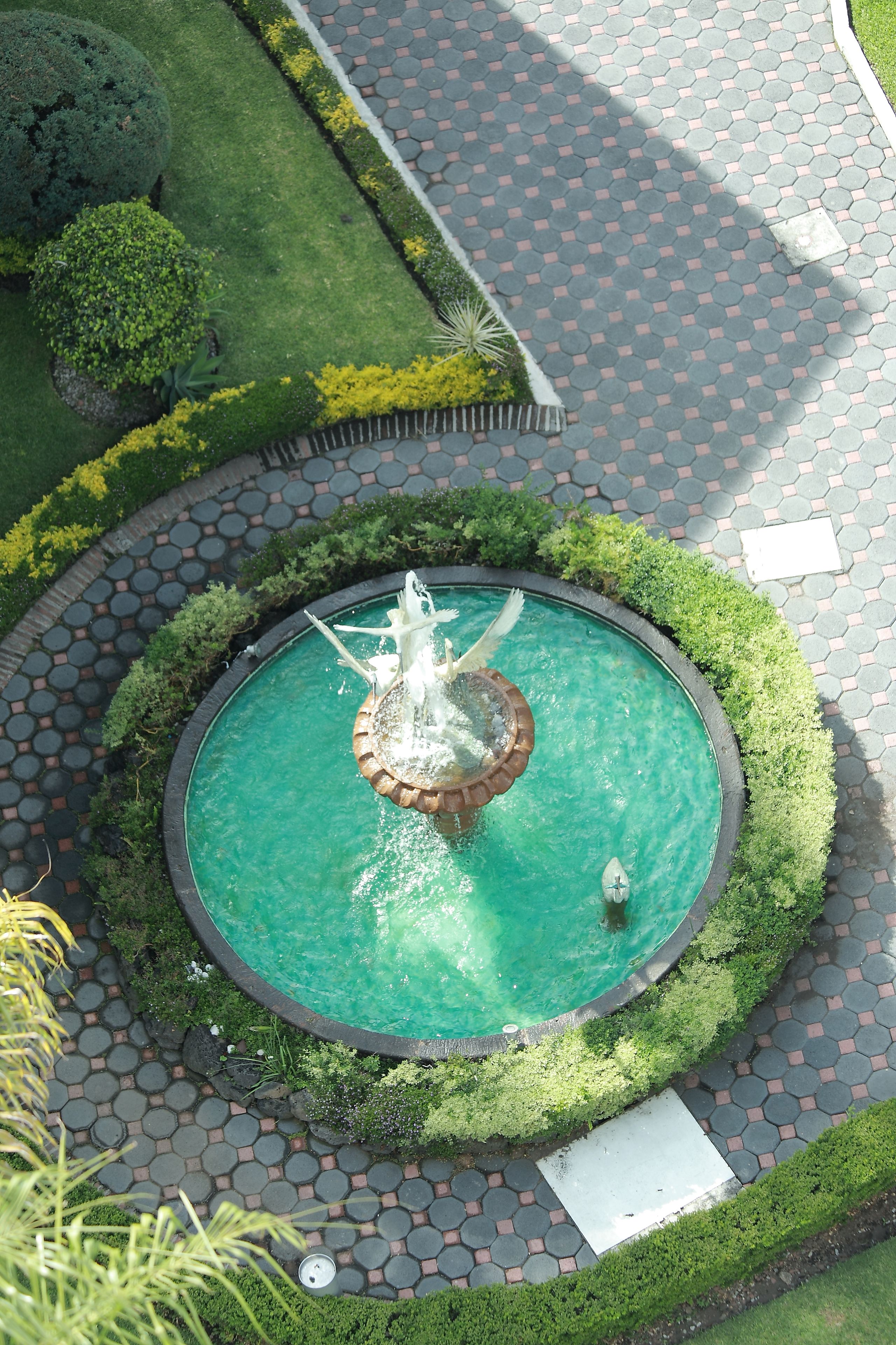 Une fontaine circulaire, ornée d’une statue centrale, est entourée d’une allée pavée.