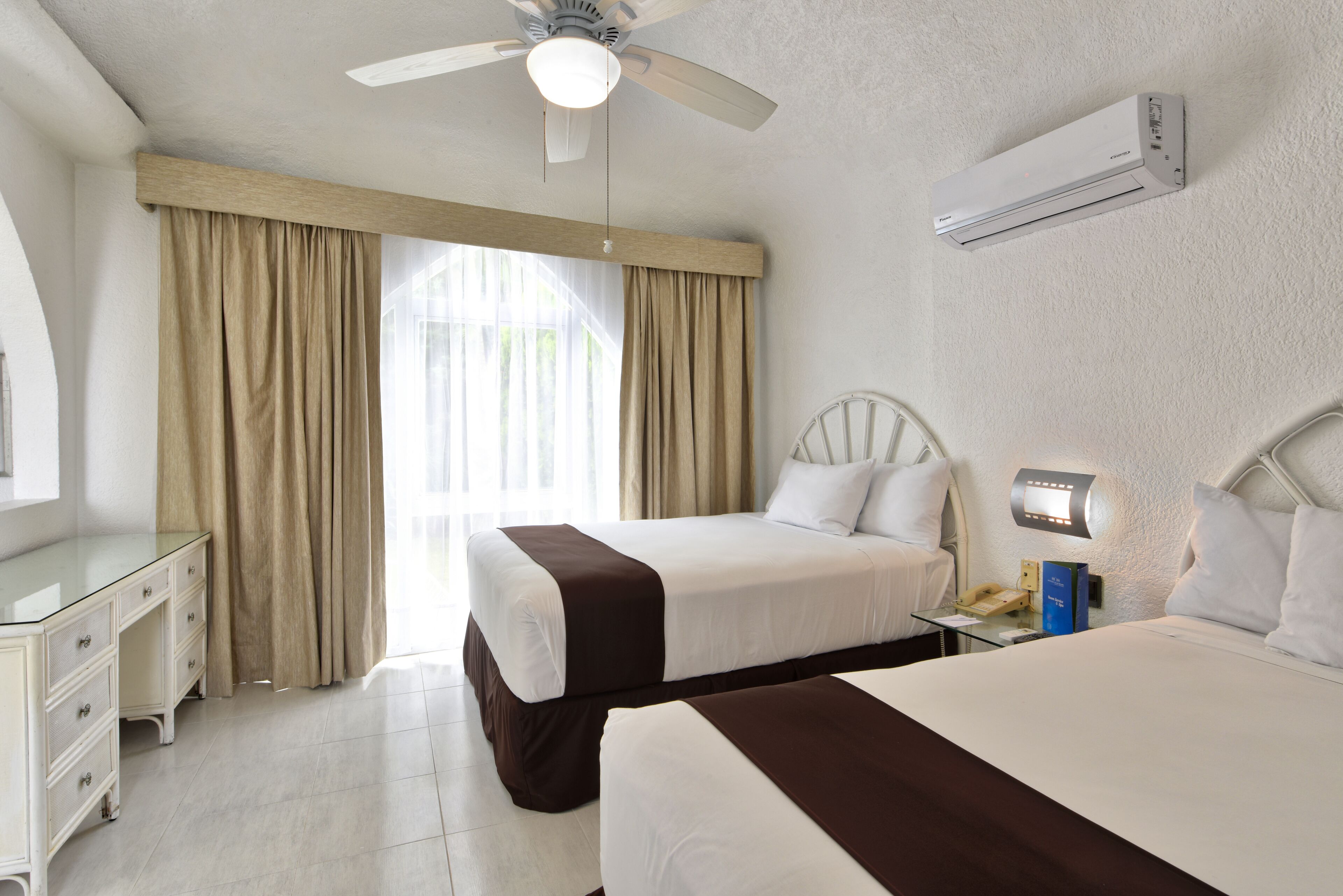 A hotel room with two beds, a desk, a ceiling fan, an air conditioner, and a bedside table with a lamp.