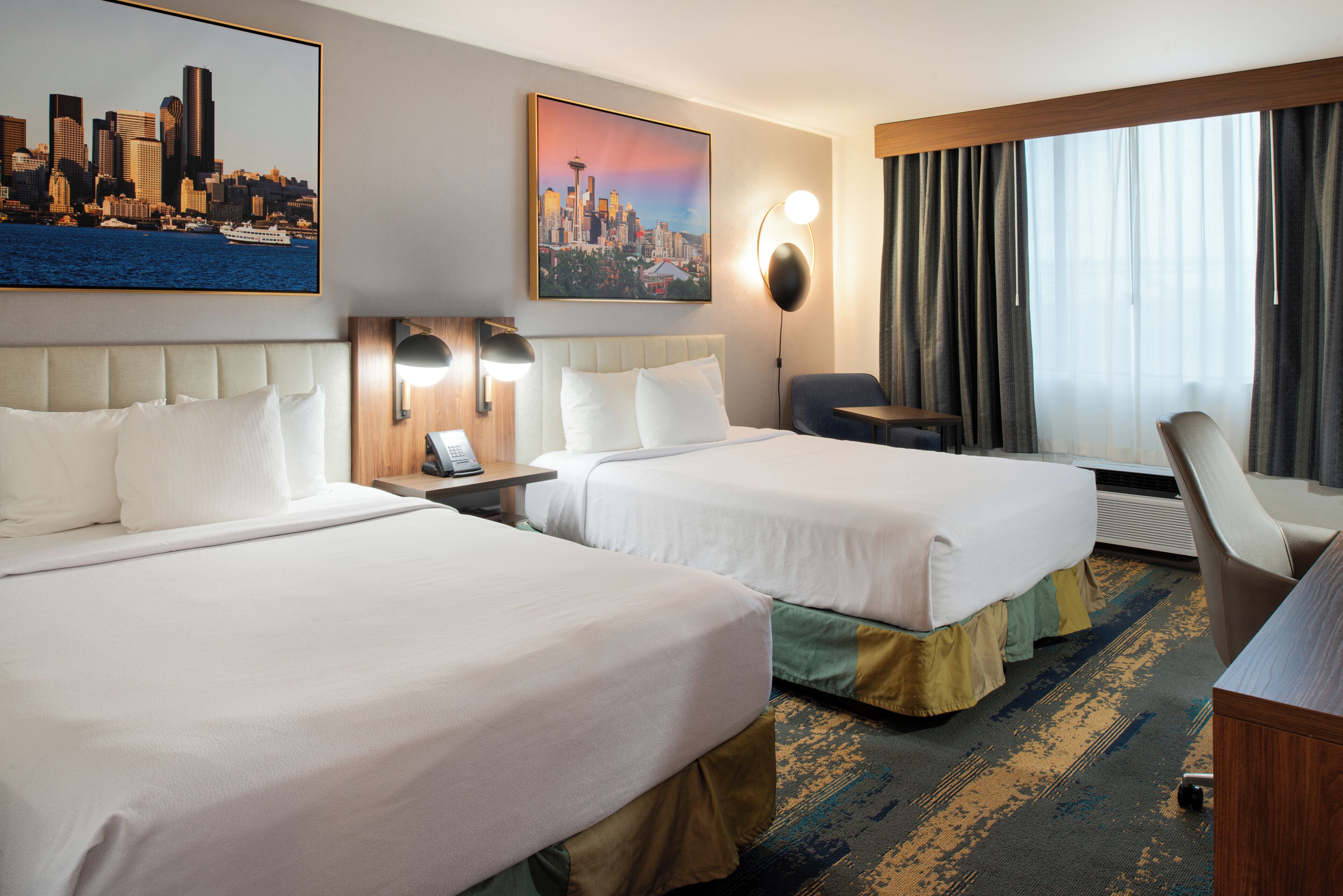 A hotel room with two beds, a desk, a chair, a cityscape painting, and a large window with curtains.