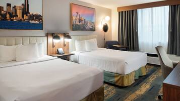 A hotel room with two beds, a desk, a chair, a cityscape painting, and a large window with curtains.