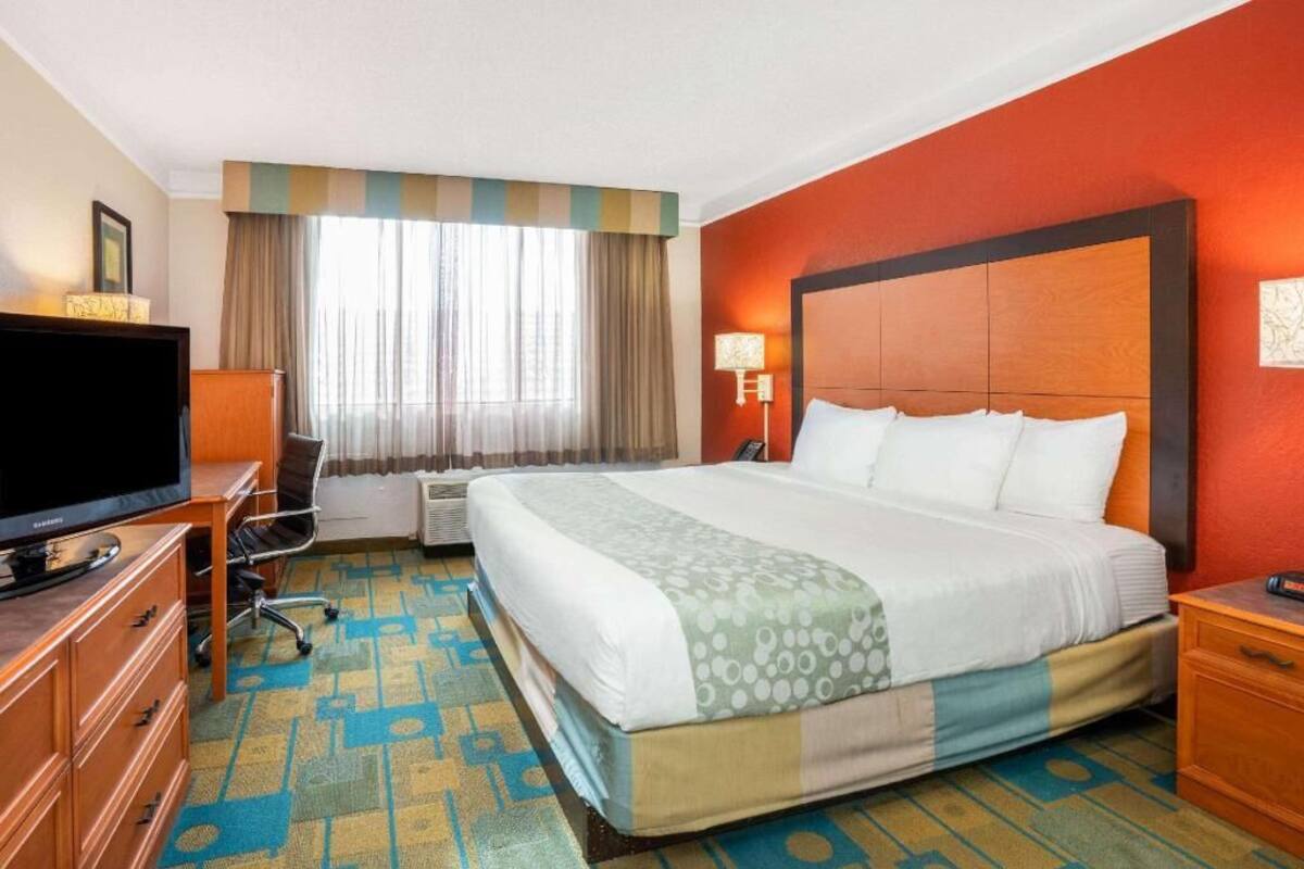 Ein Hotelzimmer mit einem großen Bett, einem Fernseher, einem Schreibtisch und einem Fenster mit Vorhängen.