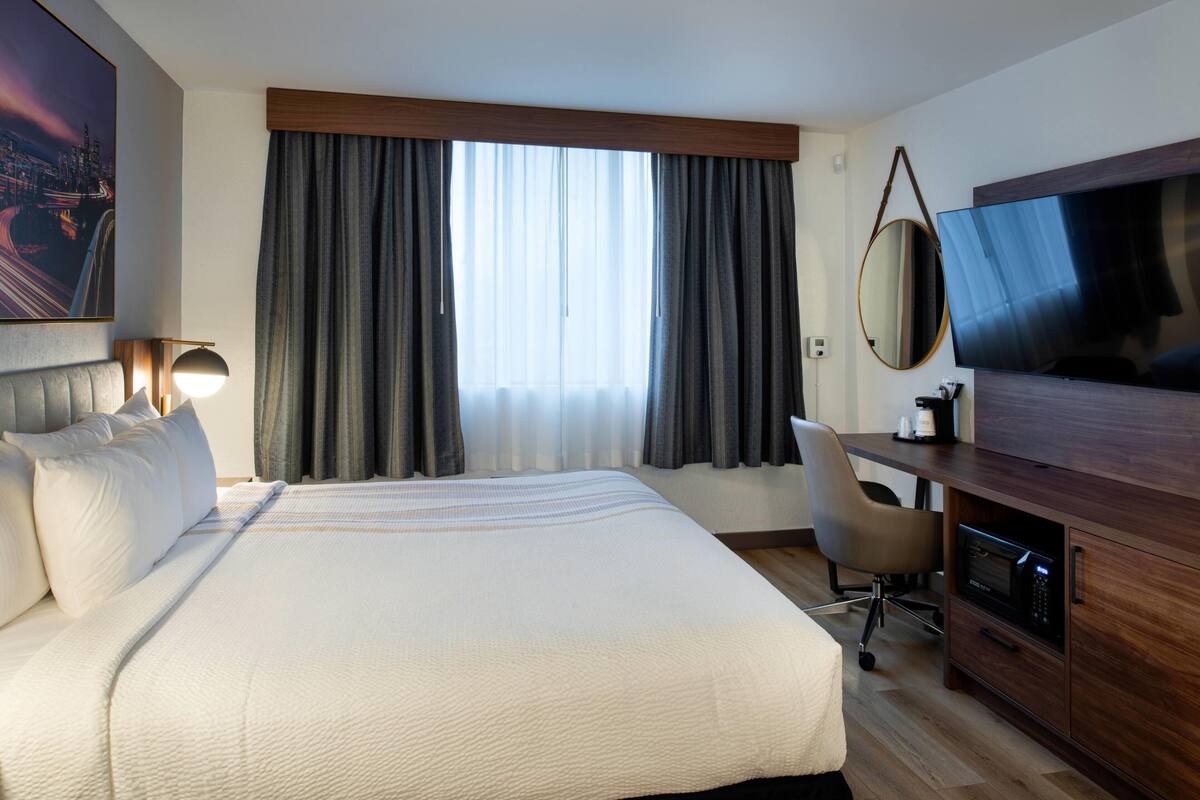 Ein Hotelzimmer mit einem großen Bett, einem Schreibtisch mit Computer, einem Fernseher und einem Fenster mit Vorhängen.