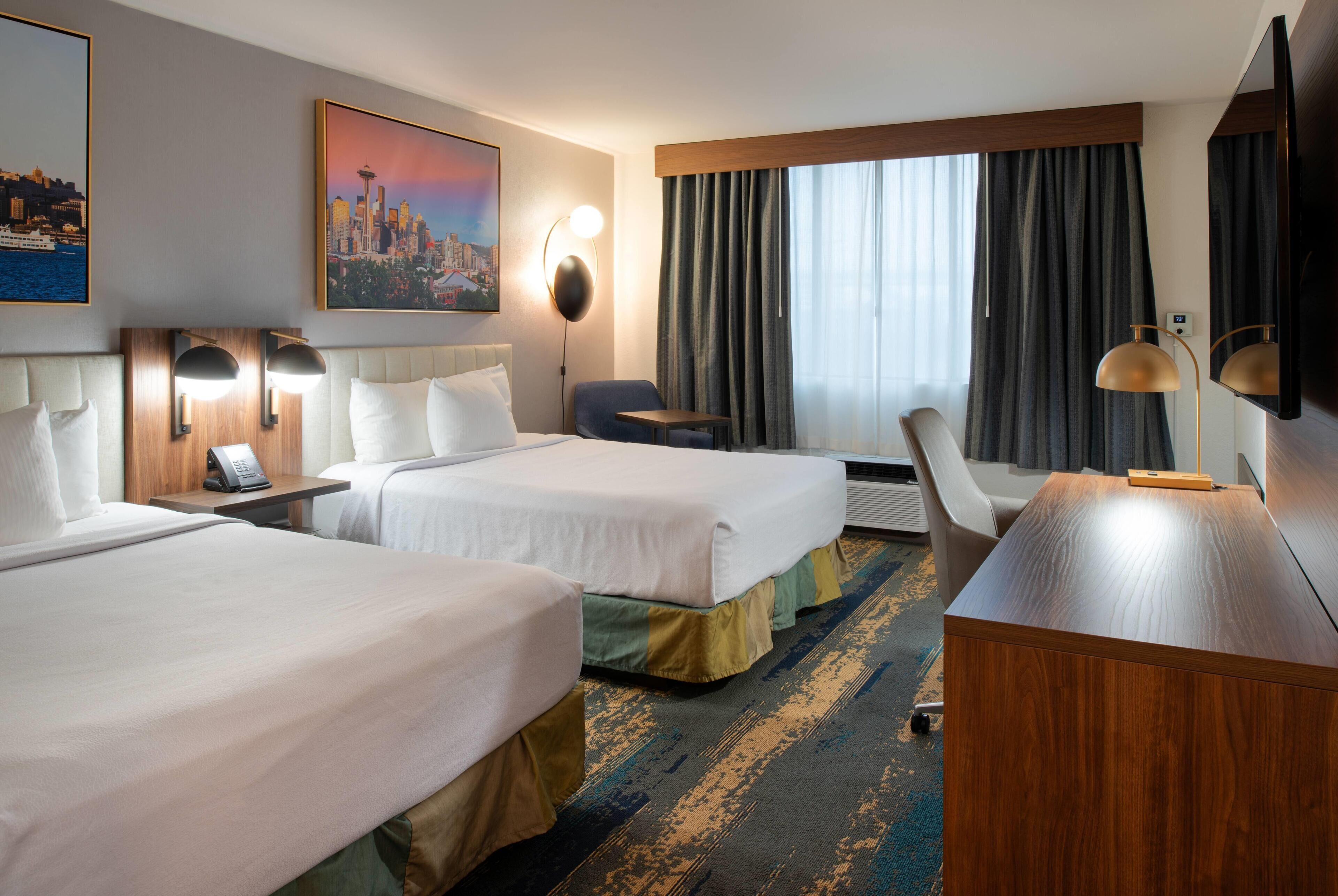 A hotel room with two beds, a desk, a chair, a phone, and a painting of a cityscape.