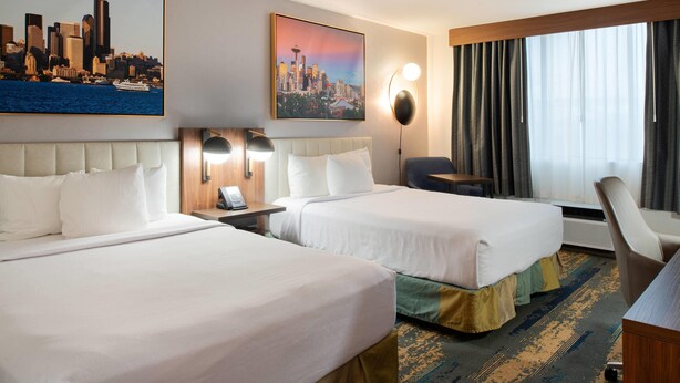 A hotel room with two beds, a desk, a chair, a large window with curtains, and two wall-mounted lamps.