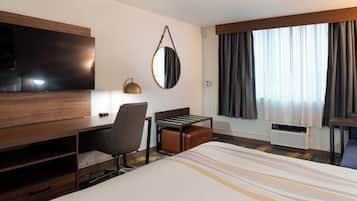 A hotel room with a bed, a desk, a chair, a TV, a mirror, a window with curtains, and an air conditioning unit.