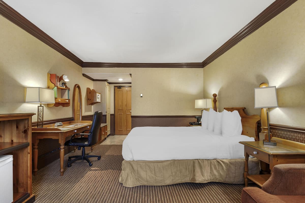 A hotel room with a large bed, a desk with a chair, a desk lamp, a chair, a nightstand, and a lamp.