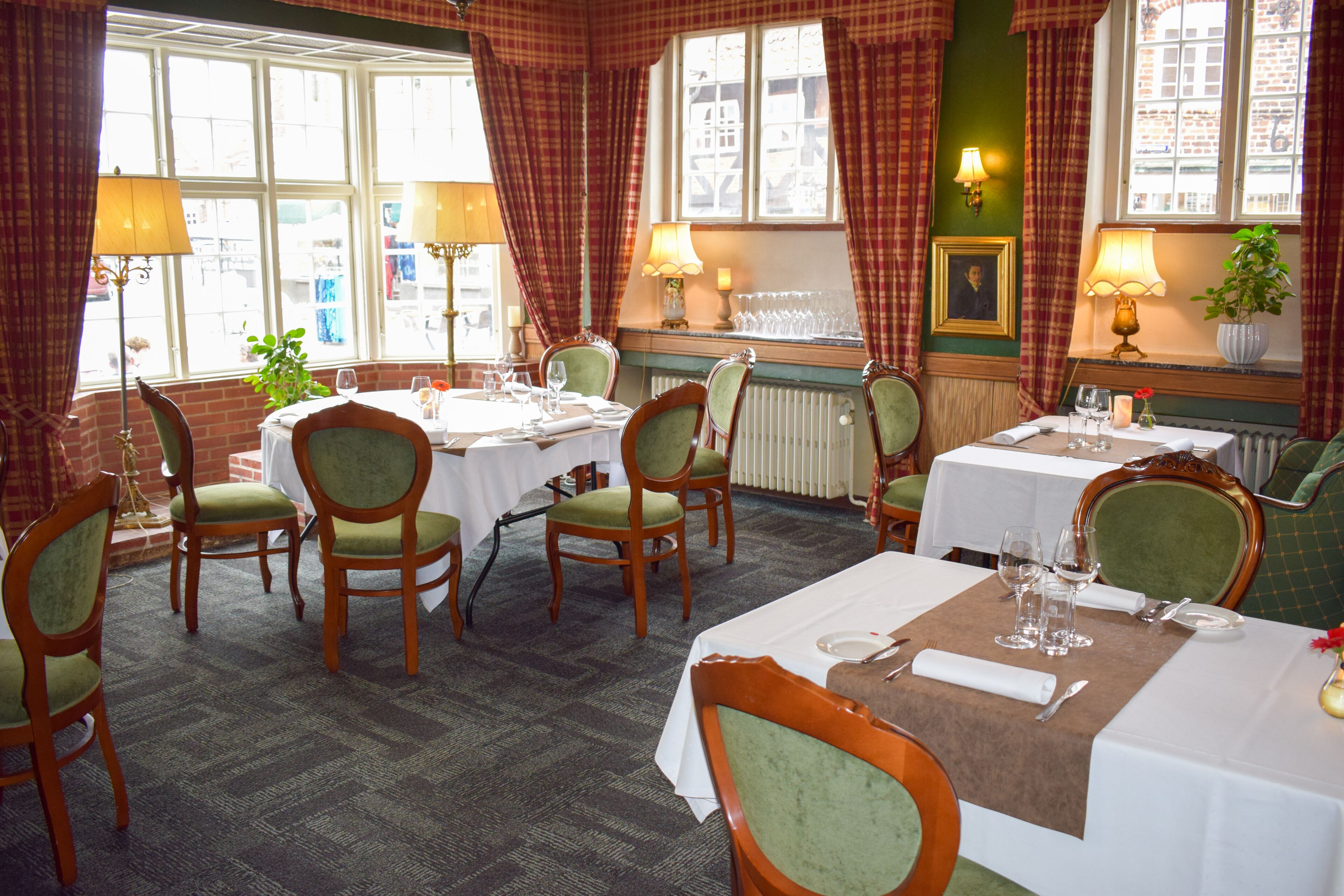 A dining area with multiple tables set for two, green upholstered chairs, and a view of the street through large windows.