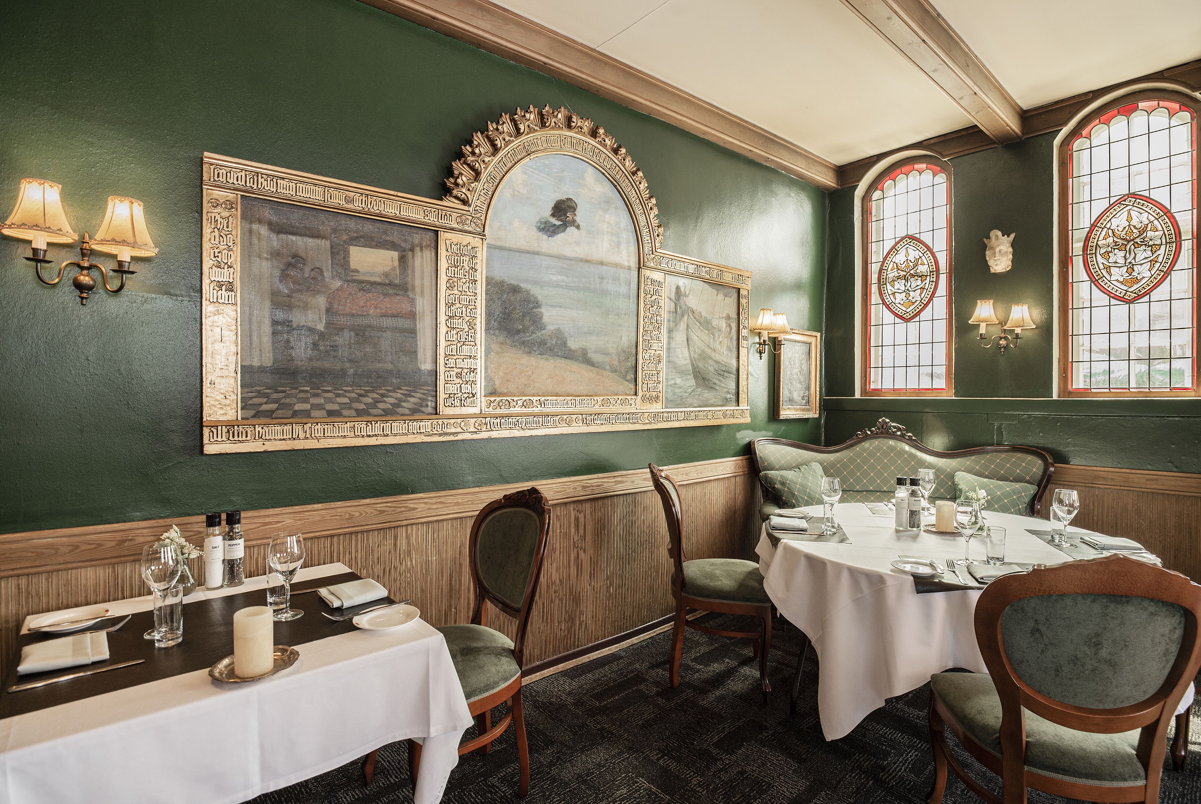 Comedor con una mesa lista para dos personas, sillas de madera, pared verde con cuadros y luces decorativas.