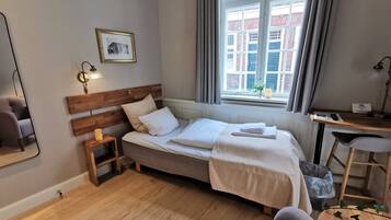 Un dormitorio ordenado con cabecera de madera, una mesita de noche con lámpara, una ventana con cortinas y vistas a edificios.