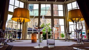 Mesa posta para refeição com taças de vinho e castiçal, grandes janelas com vista para edifícios e um candelabro.