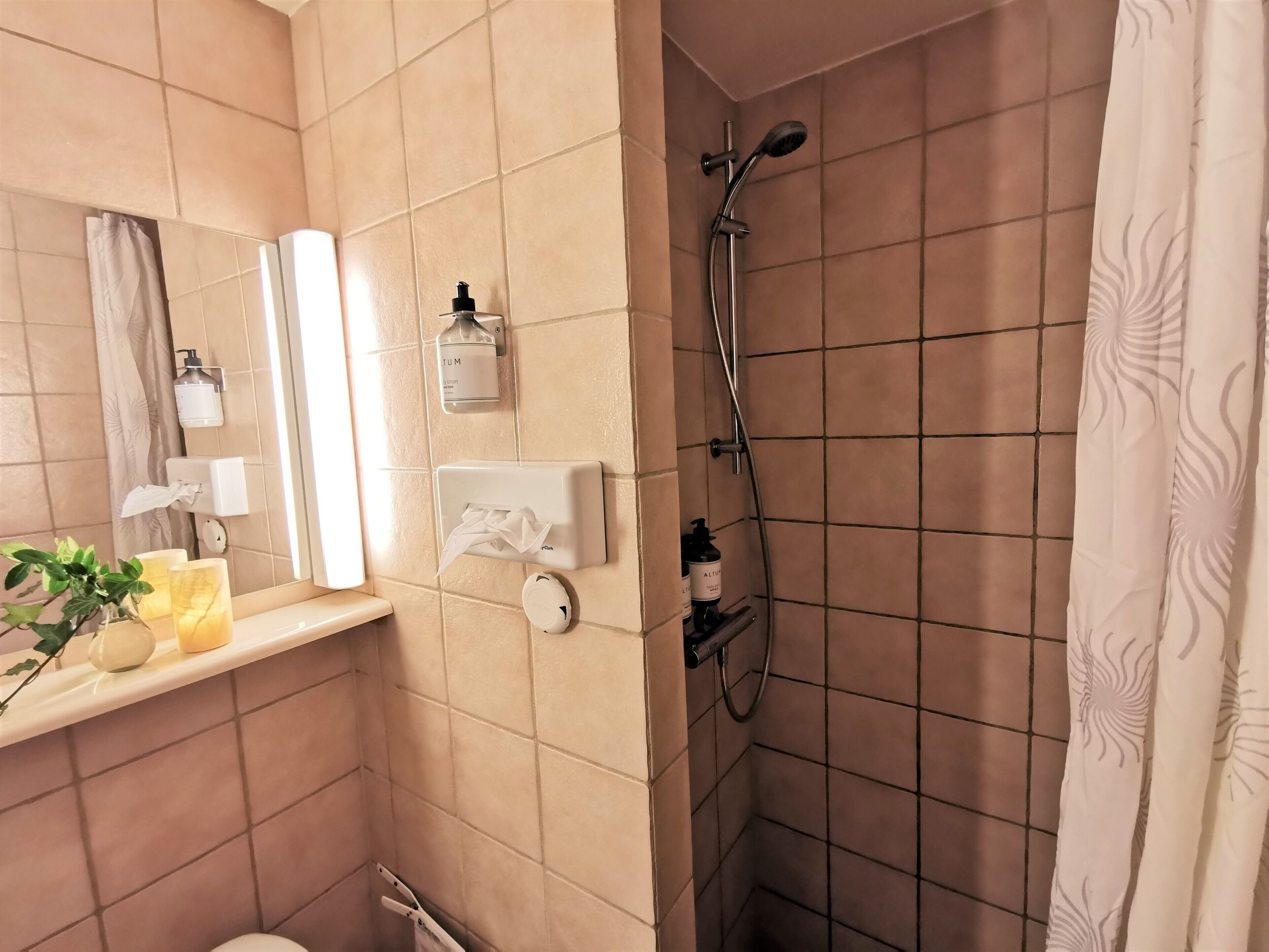 A bathroom with a mirror, towel dispenser, hand soap, and a shower.