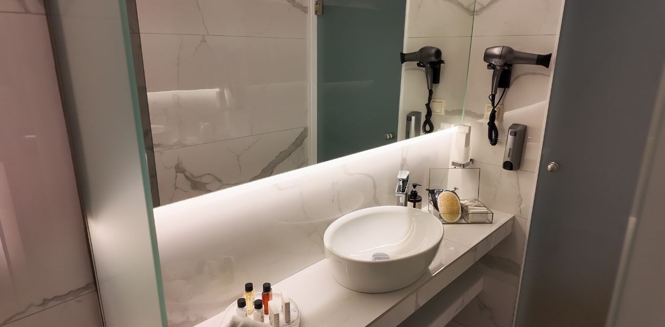 Ein modernes Badezimmer mit einem großen Spiegel, einem weißen Waschbecken und zwei an der Wand montierten Haartrocknern.