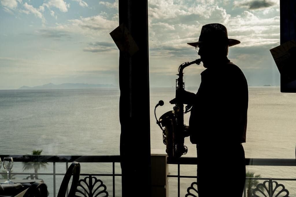 A silhouette of a person playing a saxophone on a balcony with a view of the sea and mountains.