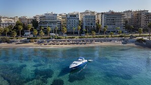 A coastal cityscape with clear blue waters, a boat, and a row of multi-story buildings. - Coral Hotel Athens (Palaio Faliro)