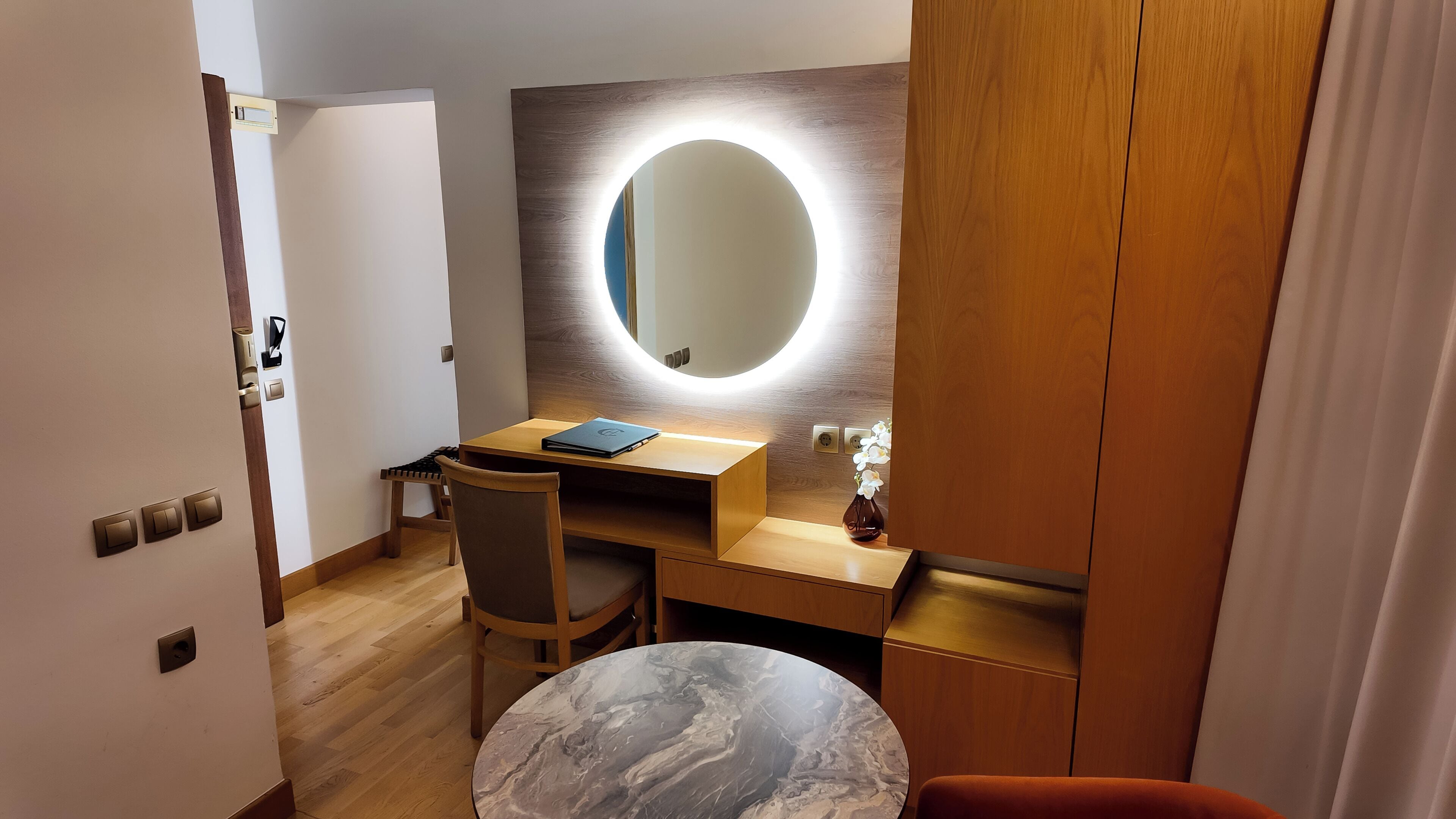 Ein kleines Zimmer mit Schreibtisch aus Holz, einem runden Spiegel mit Beleuchtung, einem kleinen Tisch und einem Stuhl.
