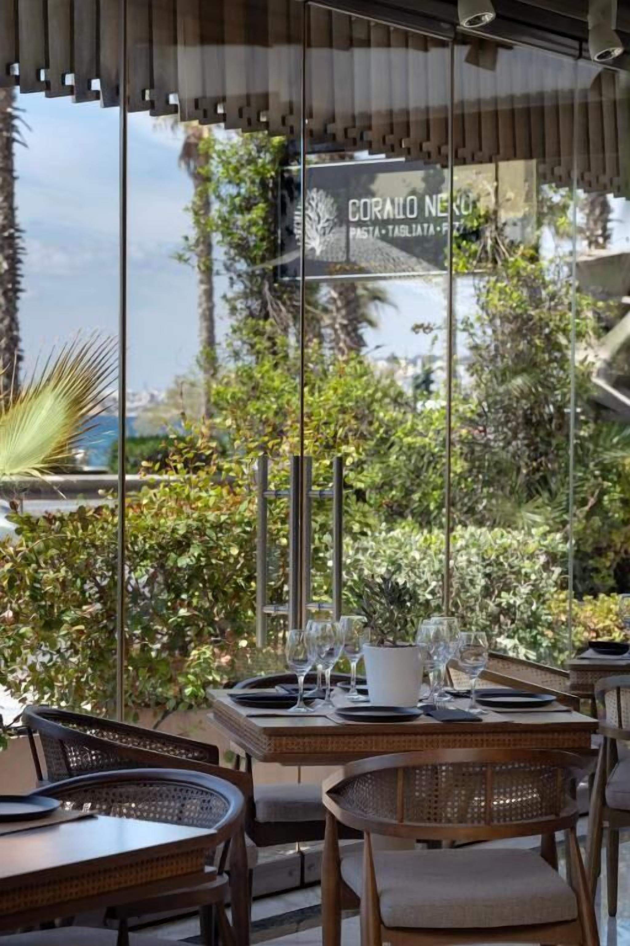 An outdoor dining area with tables and chairs, a glass wall with a sign, and lush greenery in the background.