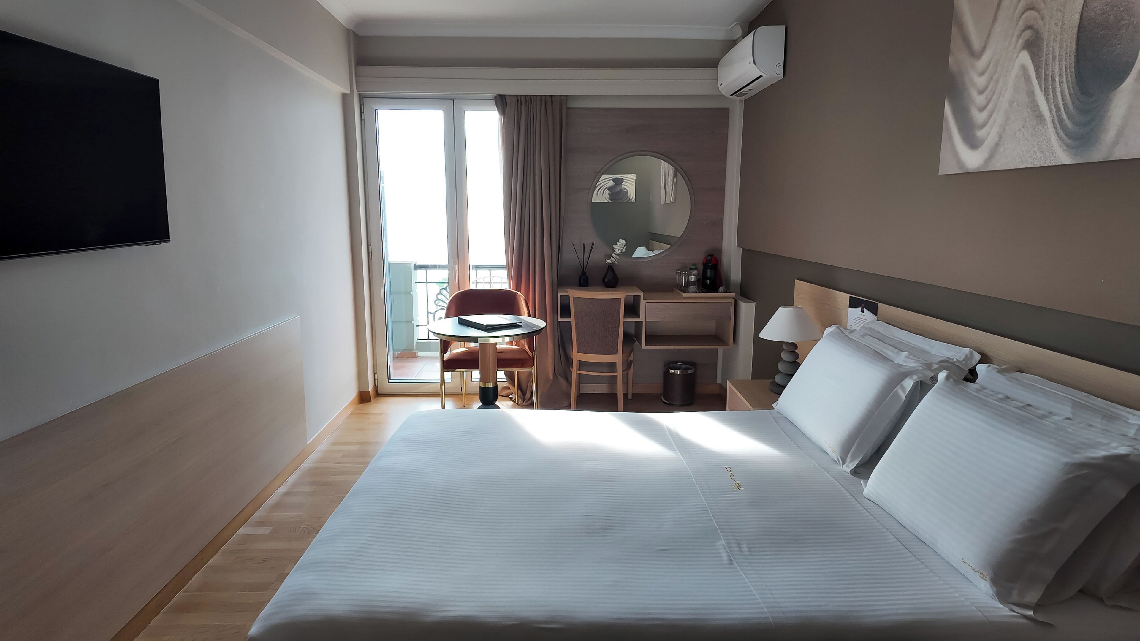A hotel room with a large bed, a desk, a chair, a TV, and a balcony with curtains.
