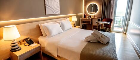 A hotel room with a large bed, bedside table with a lamp, a desk with a chair, a mirror, and a view of the ocean through a balcony.