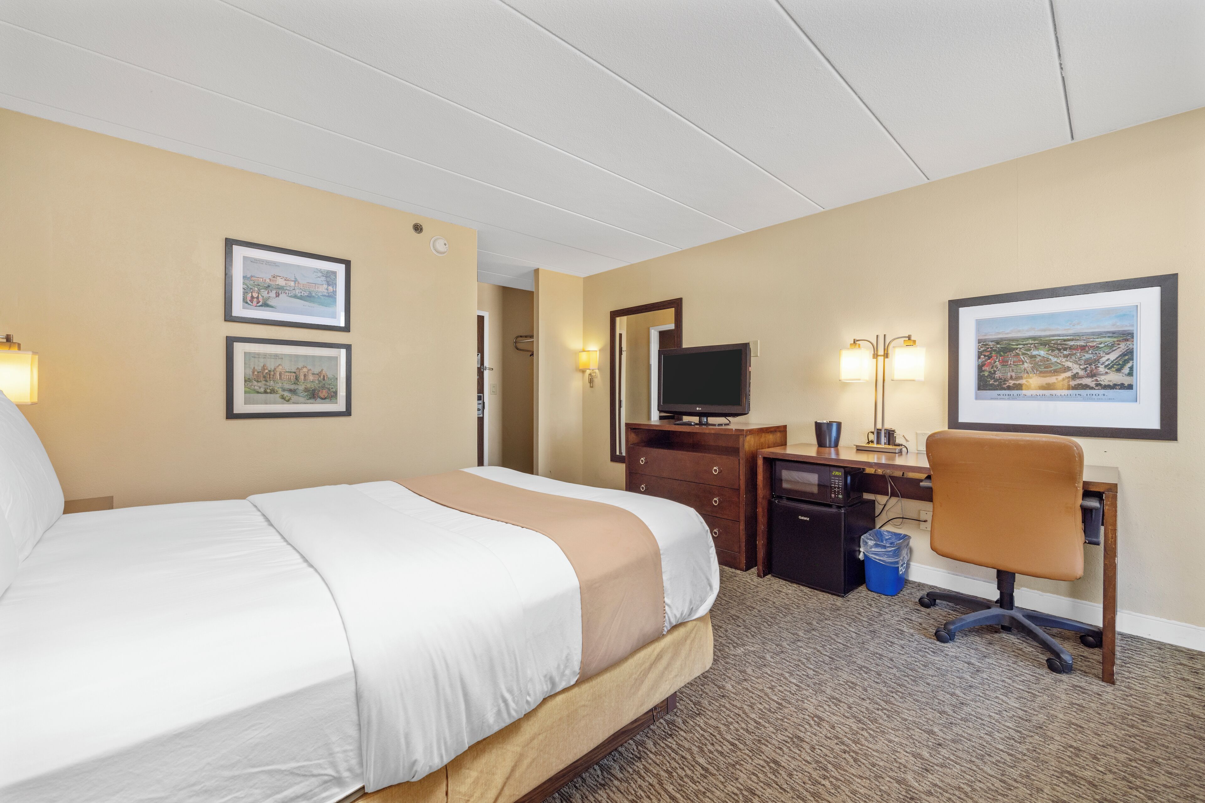 A hotel room with a bed, desk, chair, television, and framed pictures on the walls.
