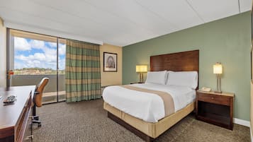 A hotel room with a large bed, two bedside tables with lamps, a desk with a chair, and a view through a large window.