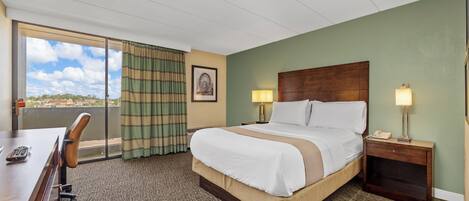 A hotel room with a large bed, two bedside tables with lamps, a desk with a chair, and a view through a large window.