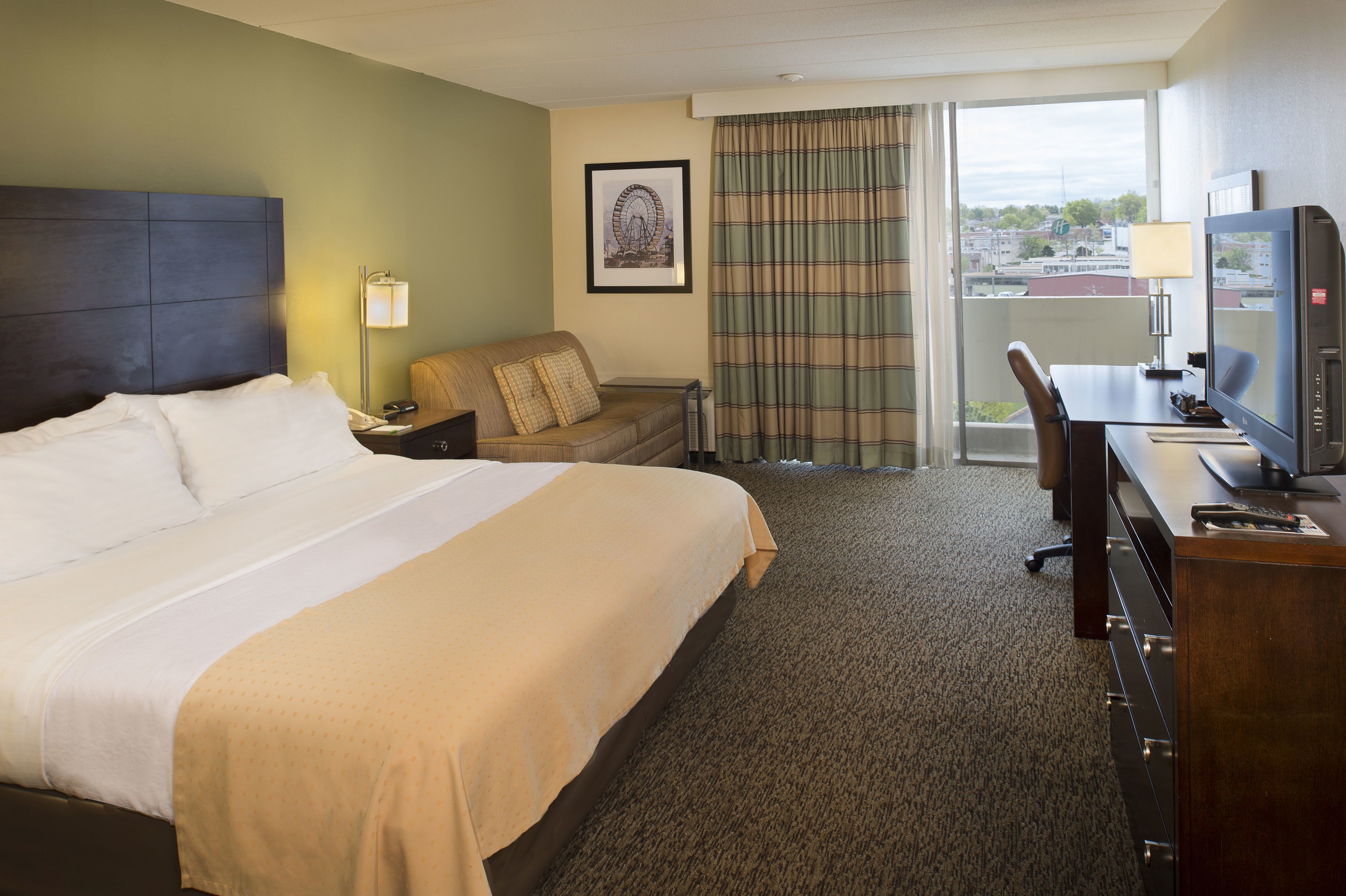 A hotel room with a large bed, a sofa, a desk with a computer, and a view of the city through the window.