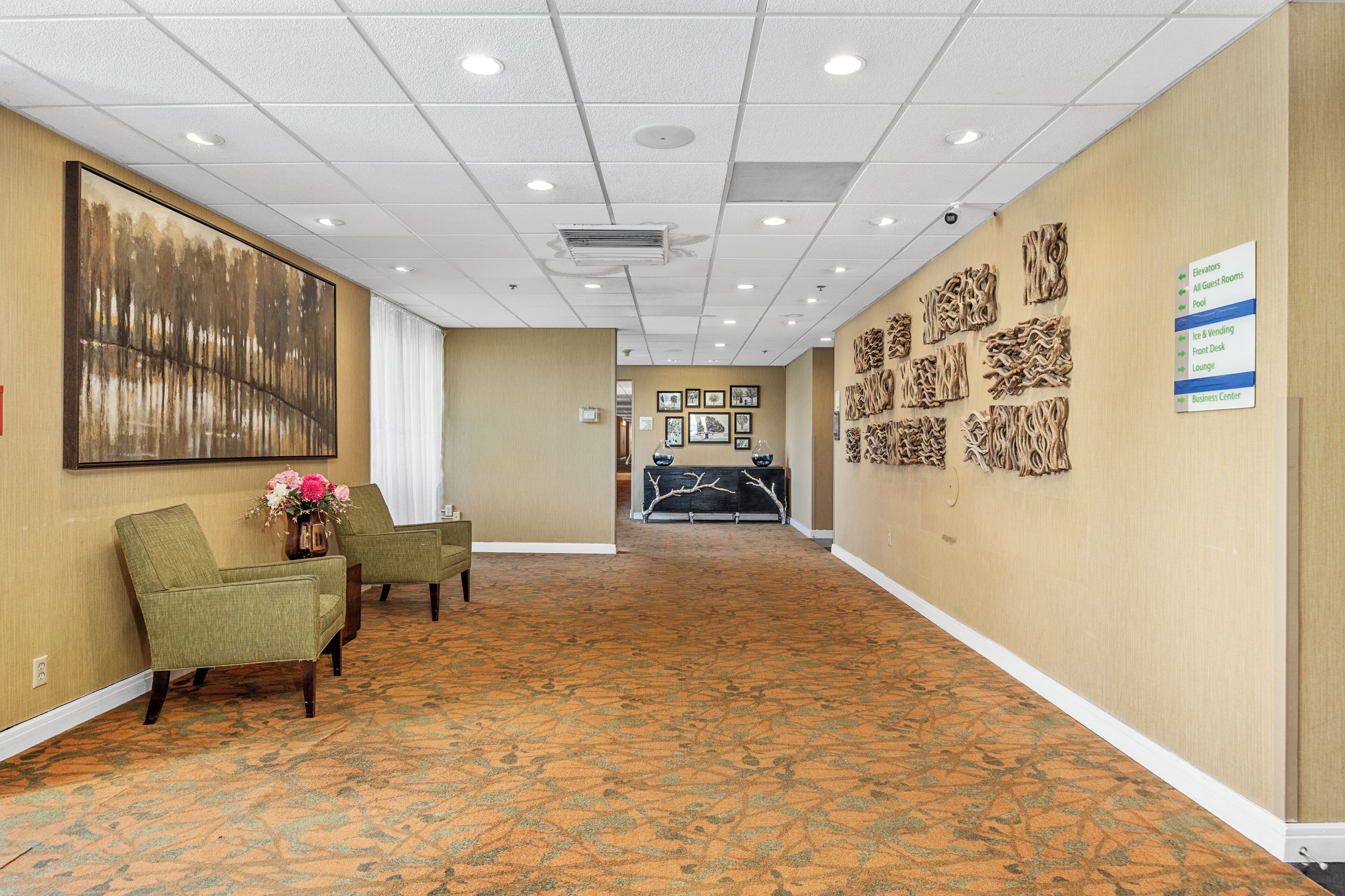A hotel corridor with a patterned carpet, green armchairs, a large abstract painting, and a sign indicating amenities like elevators, guest rooms, pool, and business center.