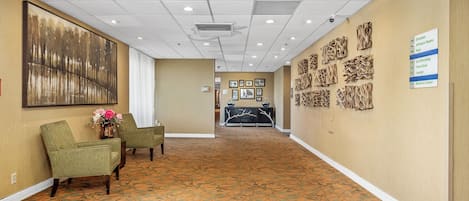 A hotel corridor with a patterned carpet, green armchairs, a large abstract painting, and a sign indicating amenities like elevators, guest rooms, pool, and business center.
