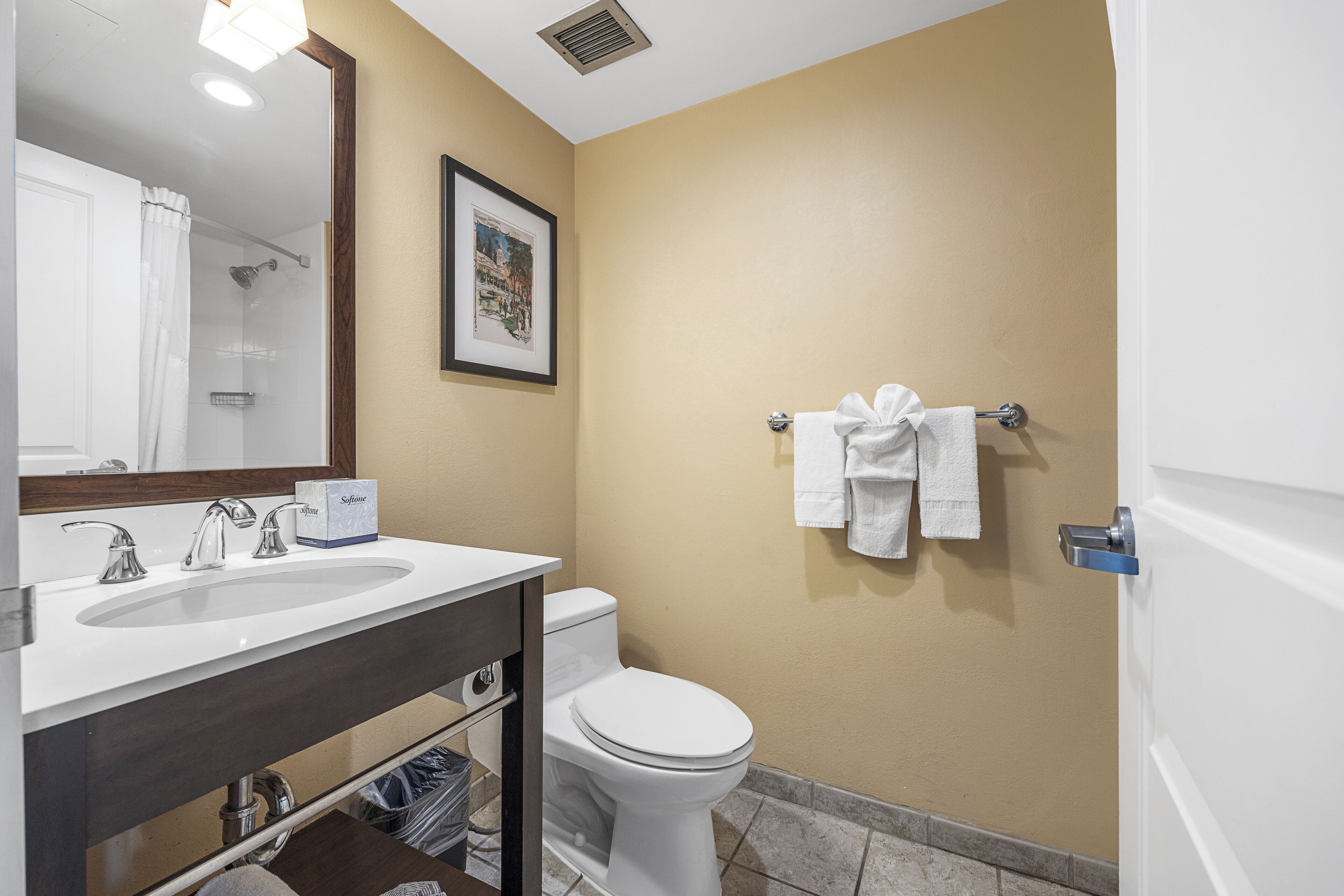 A bathroom with a white sink, a toilet, and a shower. There is a framed picture on the wall and a towel rack with towels.