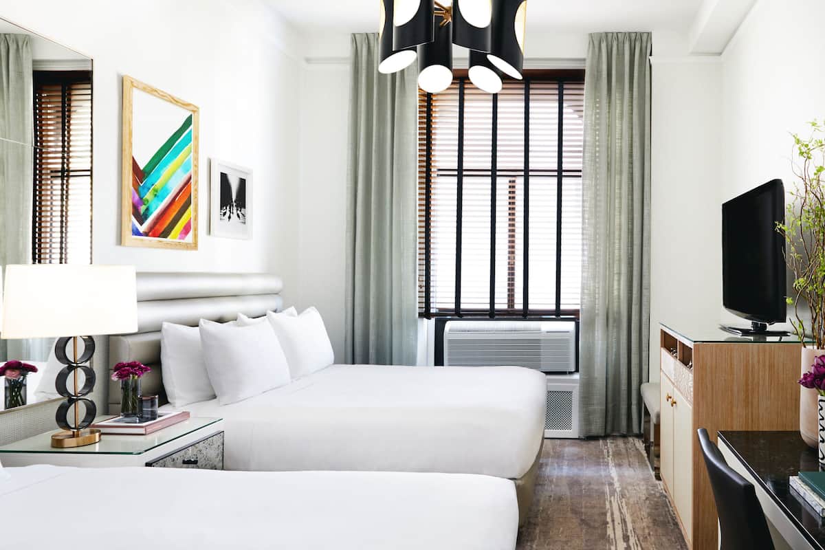 Habitación de hotel con dos camas, un escritorio, un televisor y un ventanal grande con persianas.