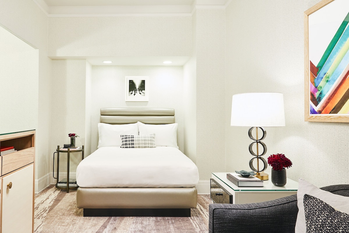 Una habitación de hotel moderna con una cama grande, una mesita de noche con lámpara, una silla y una pequeña mesa con libros y un jarrón.