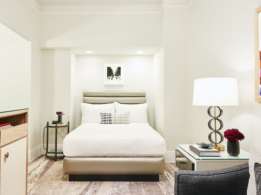A modern hotel room with a large bed, a bedside table with a lamp, a chair, and a small table with books and a vase.