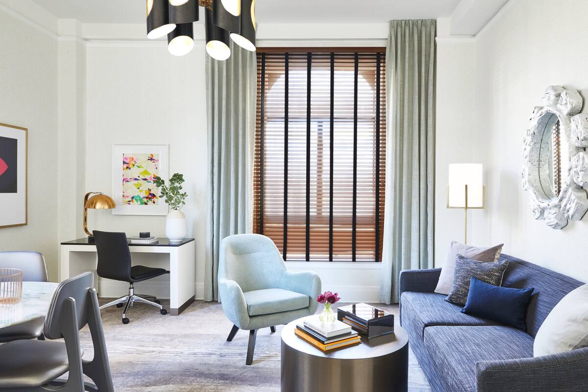 Una sala de estar moderna con un sillón azul claro, un sofá azul, una mesa de centro redonda con libros, un escritorio con una lámpara y un ventanal con persianas.