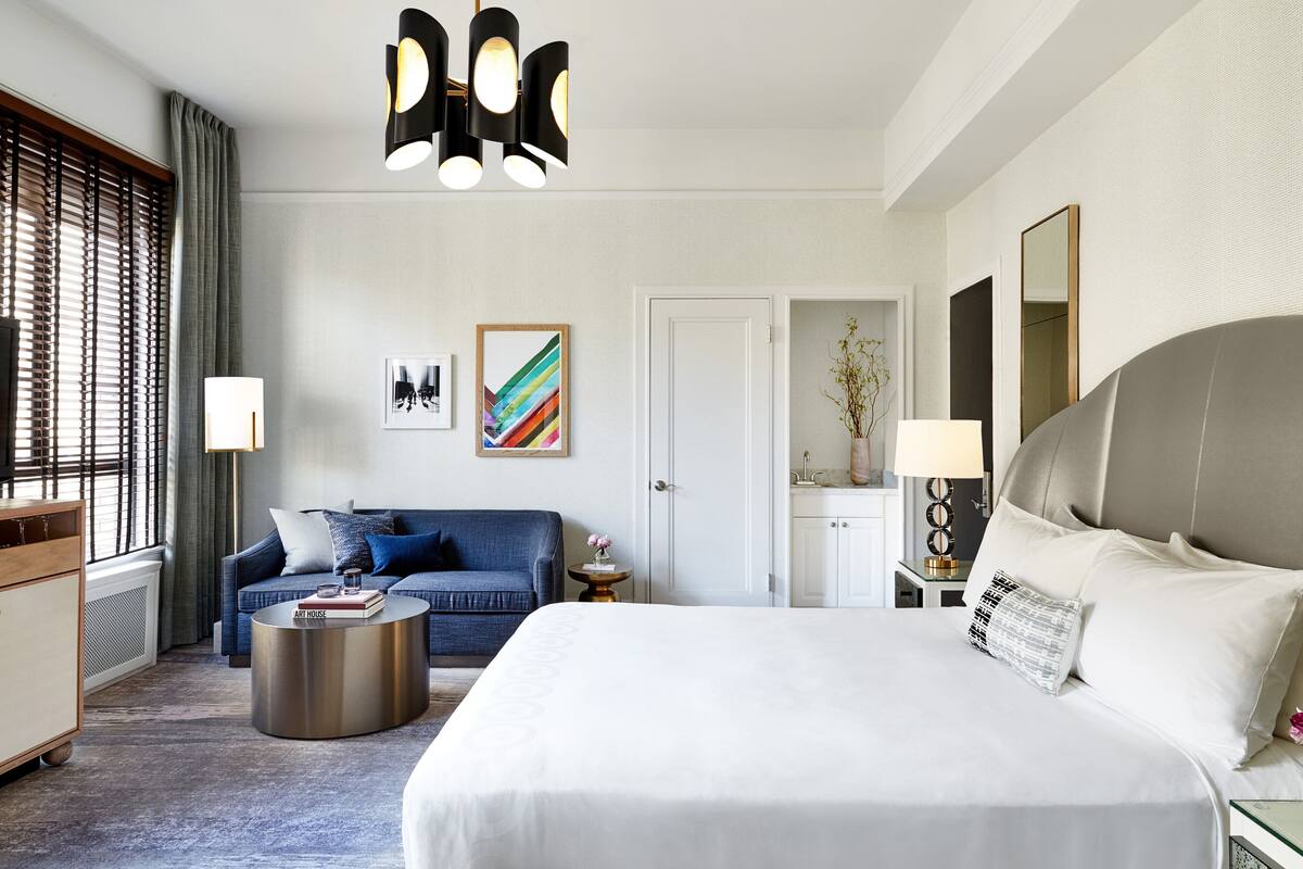Una habitación de hotel con una cama grande, un sofá, una mesa de centro redonda, un escritorio con lámpara y un espejo grande.