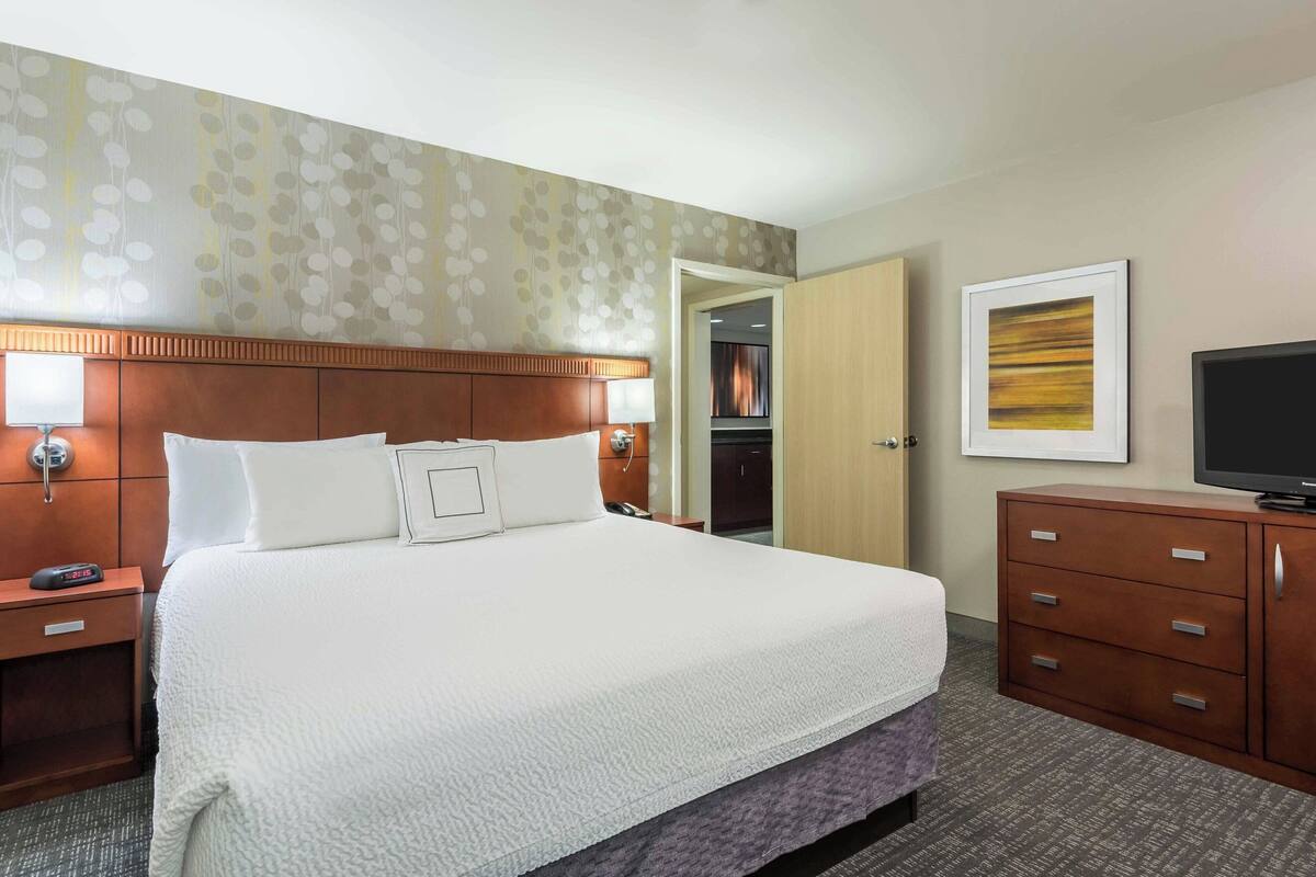 Una habitación de hotel con una cama grande, cabecera de madera, mesita de noche con lámpara, cómoda, televisor y puerta que da a un armario.