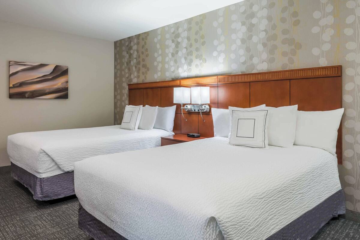 Habitación de hotel con dos camas, cabecero, mesita de noche, lámpara y pared con papel tapiz estampado.