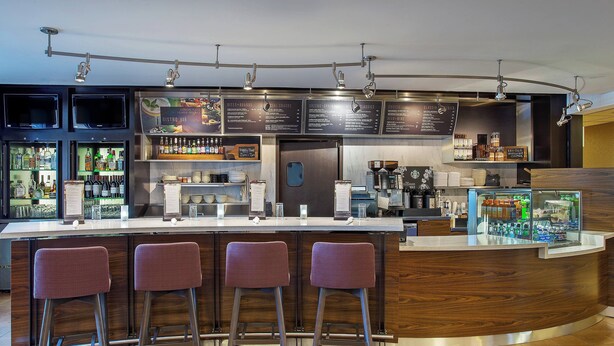 A modern bar area with a curved counter, three bar stools, and a variety of beverages and food items displayed. - Courtyard by Marriott Knoxville Cedar Bluff (Knoxville)