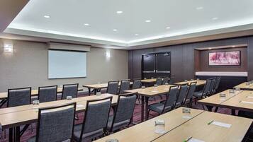 Un salón de conferencias con varias mesas y sillas, una pantalla de proyector y un cuadro grande en la pared.