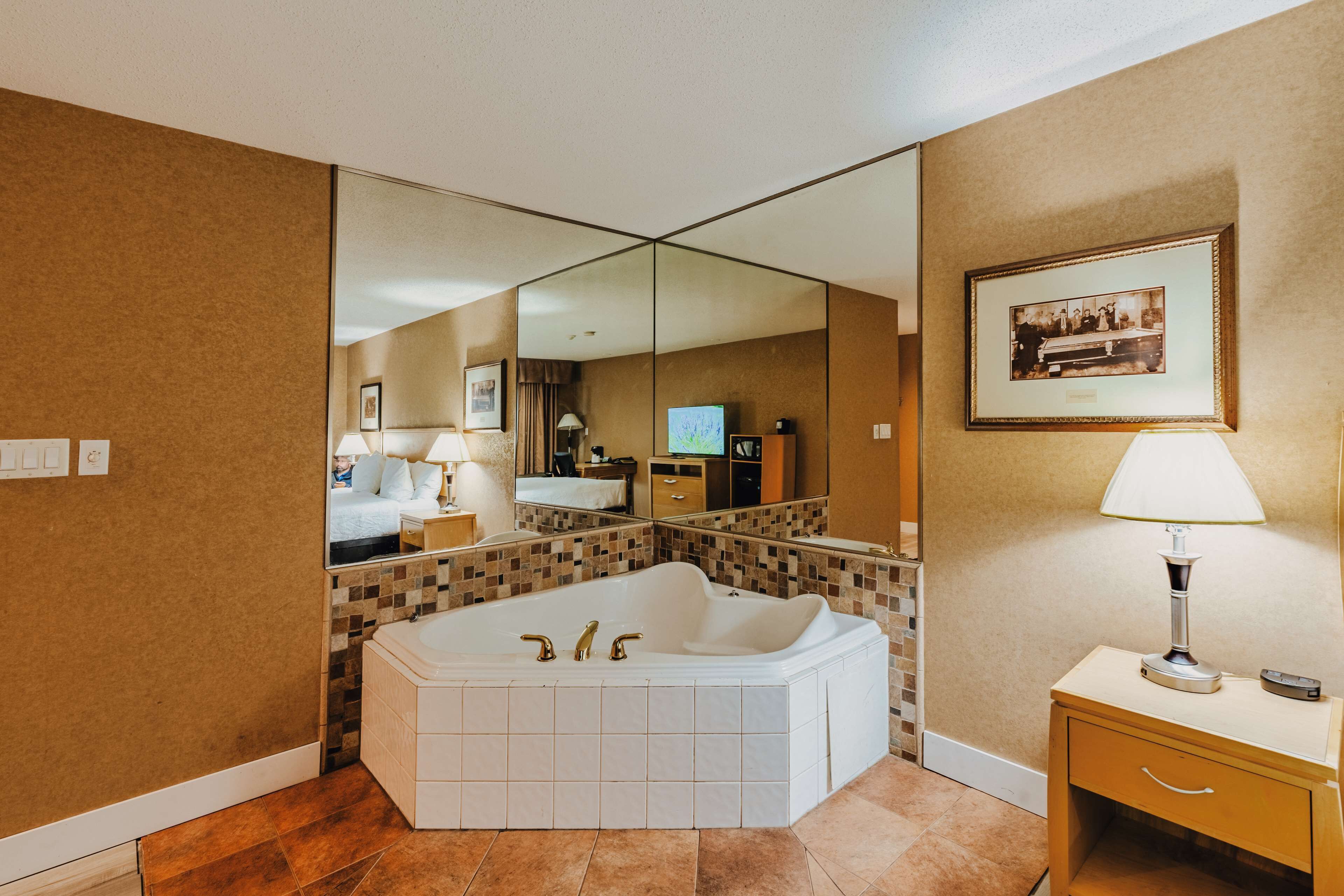 A modern bathroom with a large bathtub, a mirror, a framed picture, and a bedside table with a lamp.