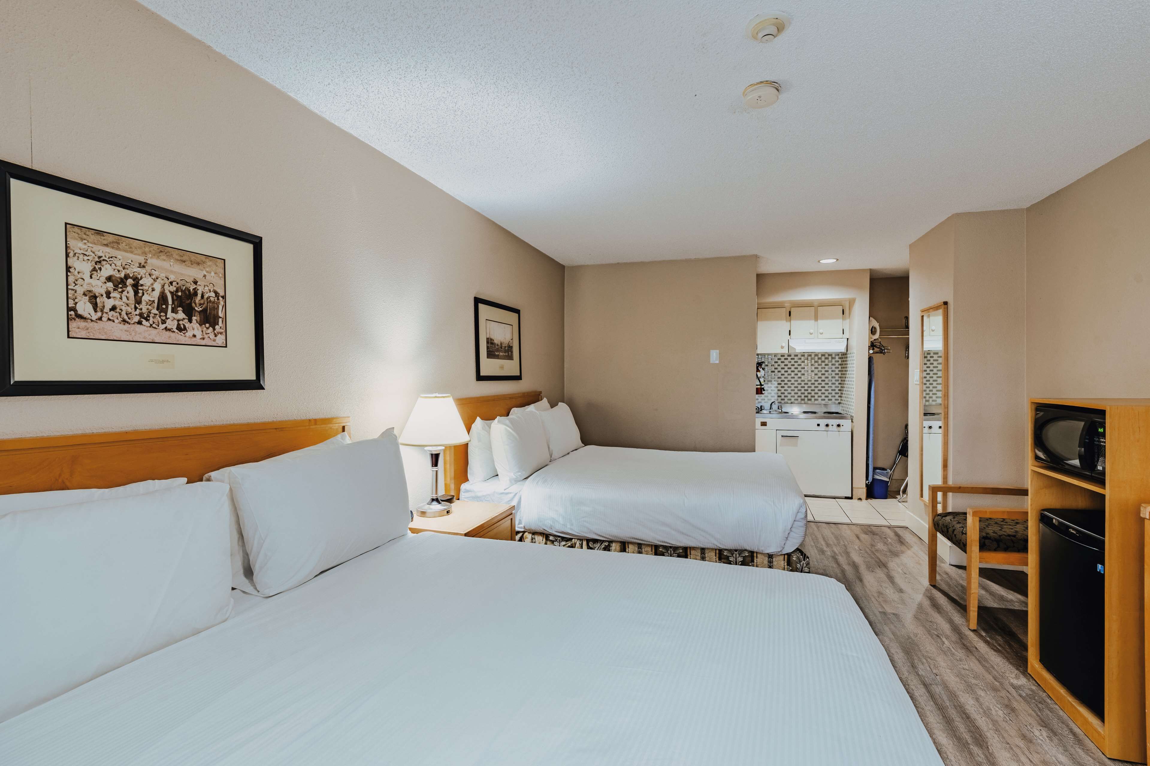 A hotel room with two beds, a desk with a lamp, a television, and a kitchenette in the background.