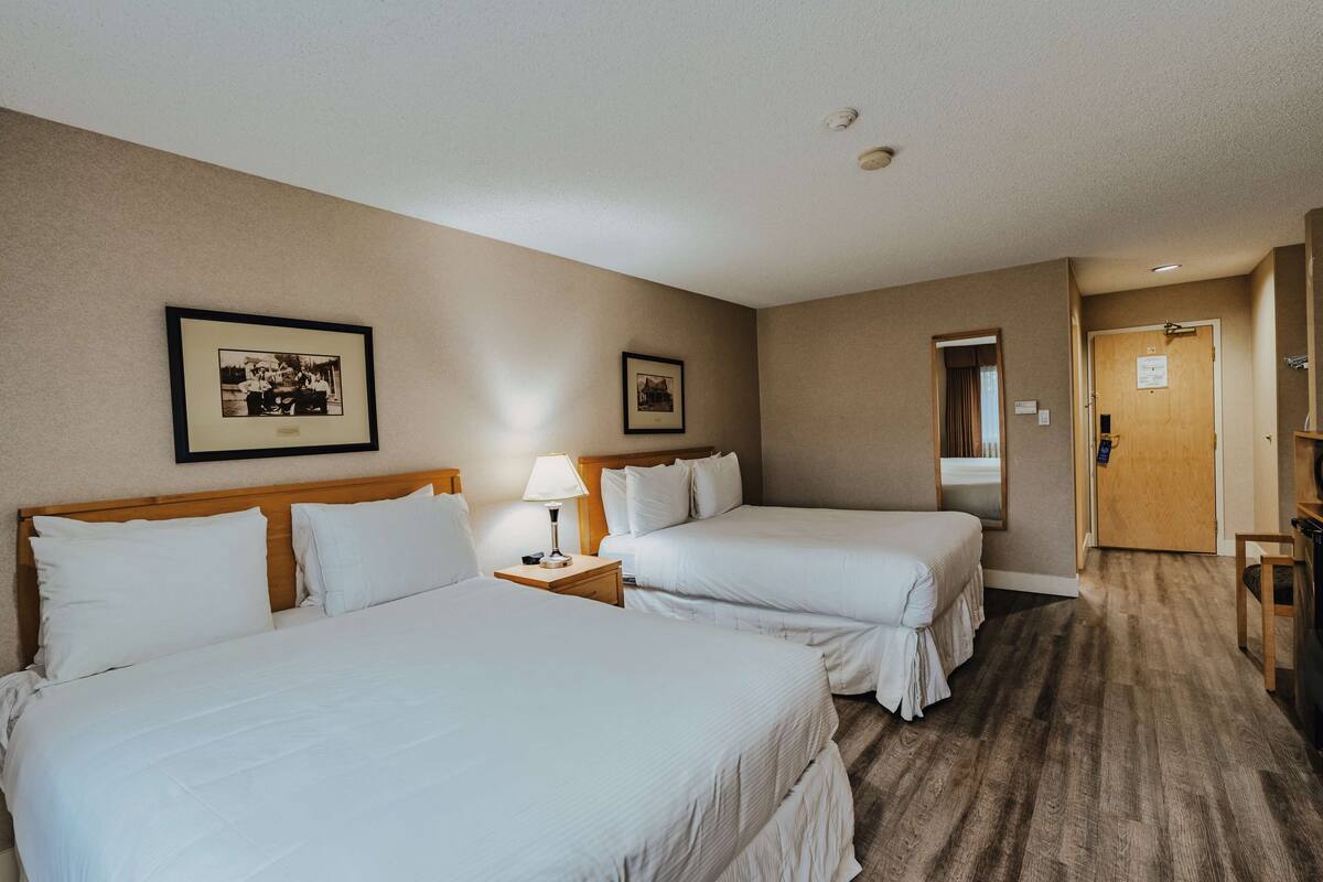Habitación de hotel con dos camas, una mesita de noche con lámpara, cuadros en la pared y una puerta de madera.