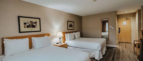 A hotel room with two beds, a nightstand with a lamp, framed pictures on the wall, and a wooden door.