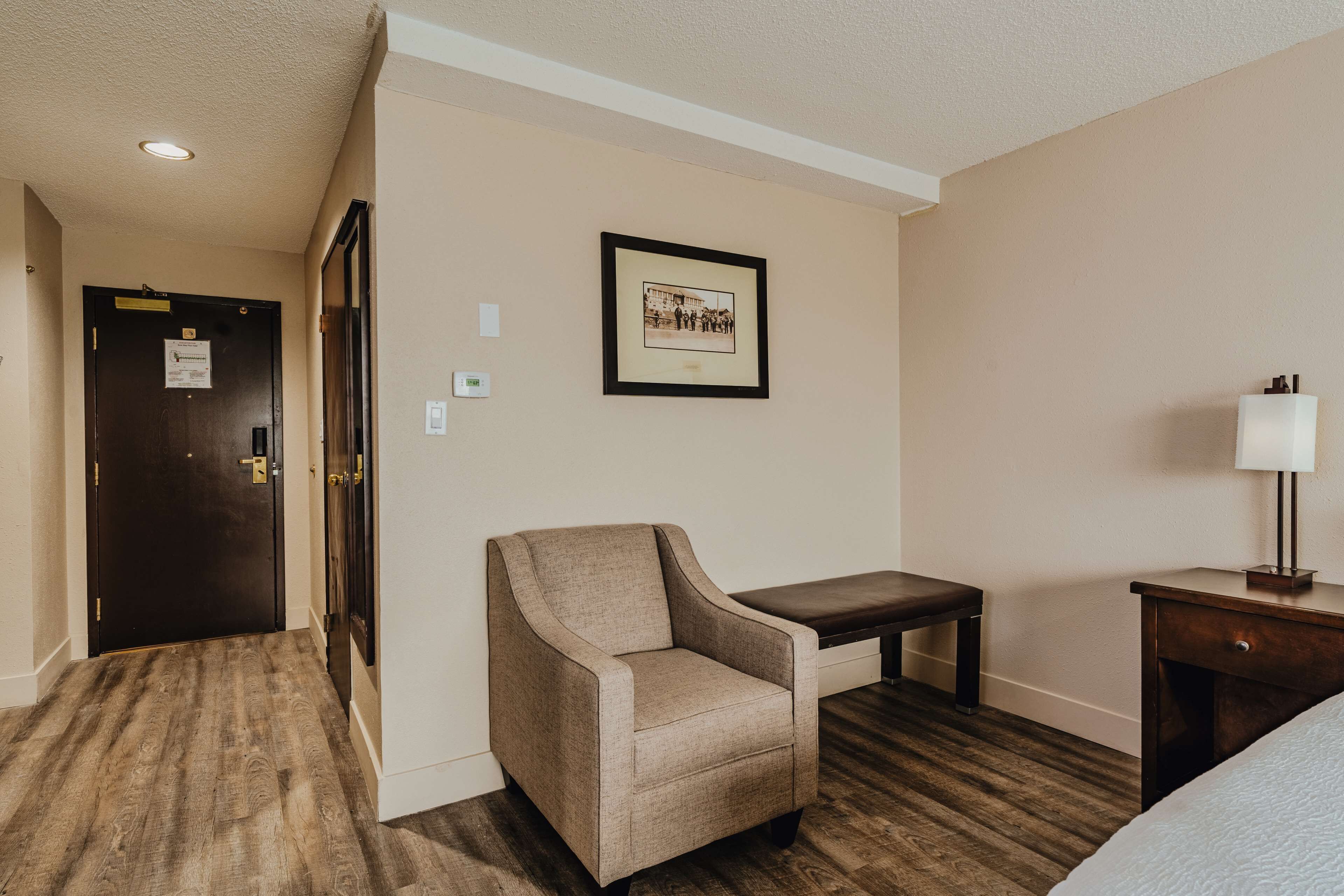 Une chambre d’hôtel avec un plancher en bois, un fauteuil beige, une petite table, un banc, une table de chevet avec une lampe et un tableau encadré au mur.