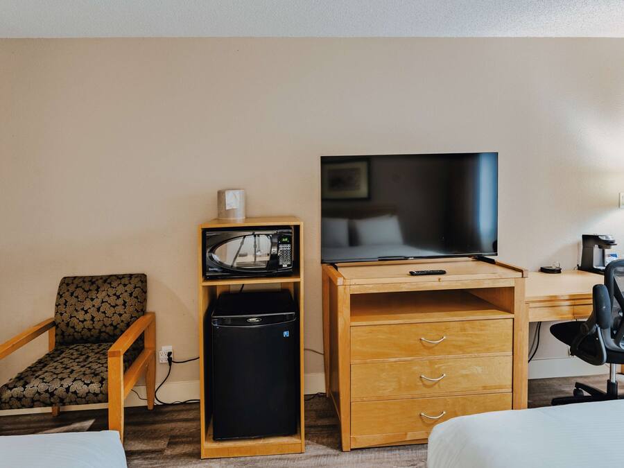 A hotel room with a bed, a desk with a flat-screen TV, a chair, a small refrigerator, and a trash bin.