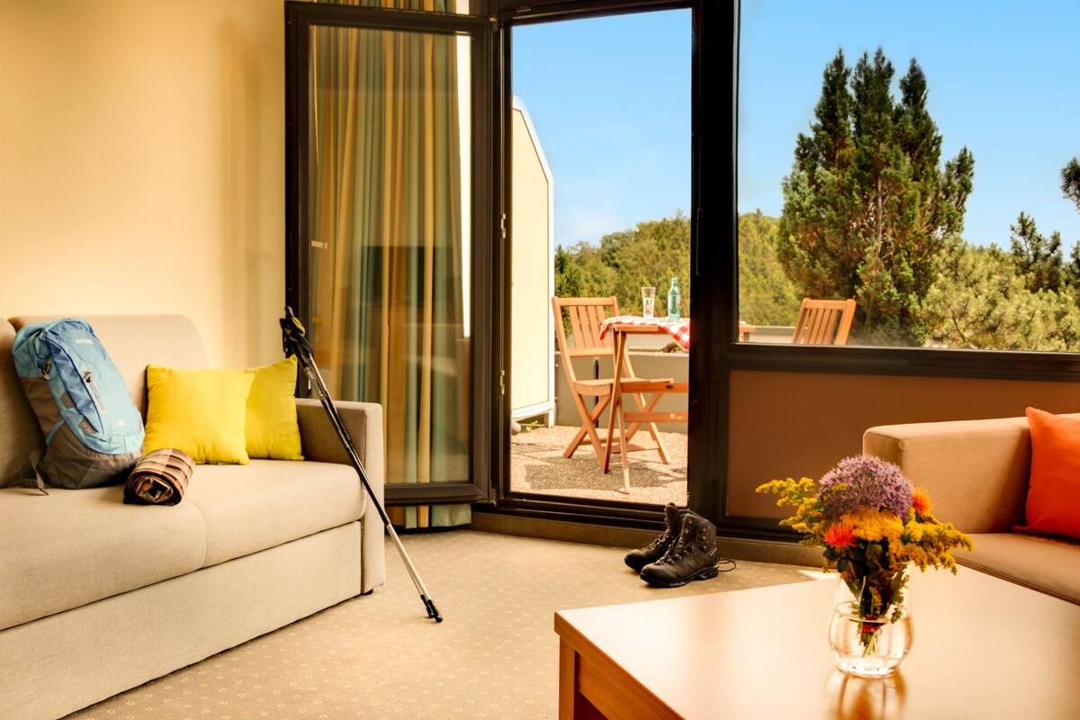 A room with a sofa, a coffee table, a vase with flowers, a pair of boots, a suitcase, and a wheelchair.