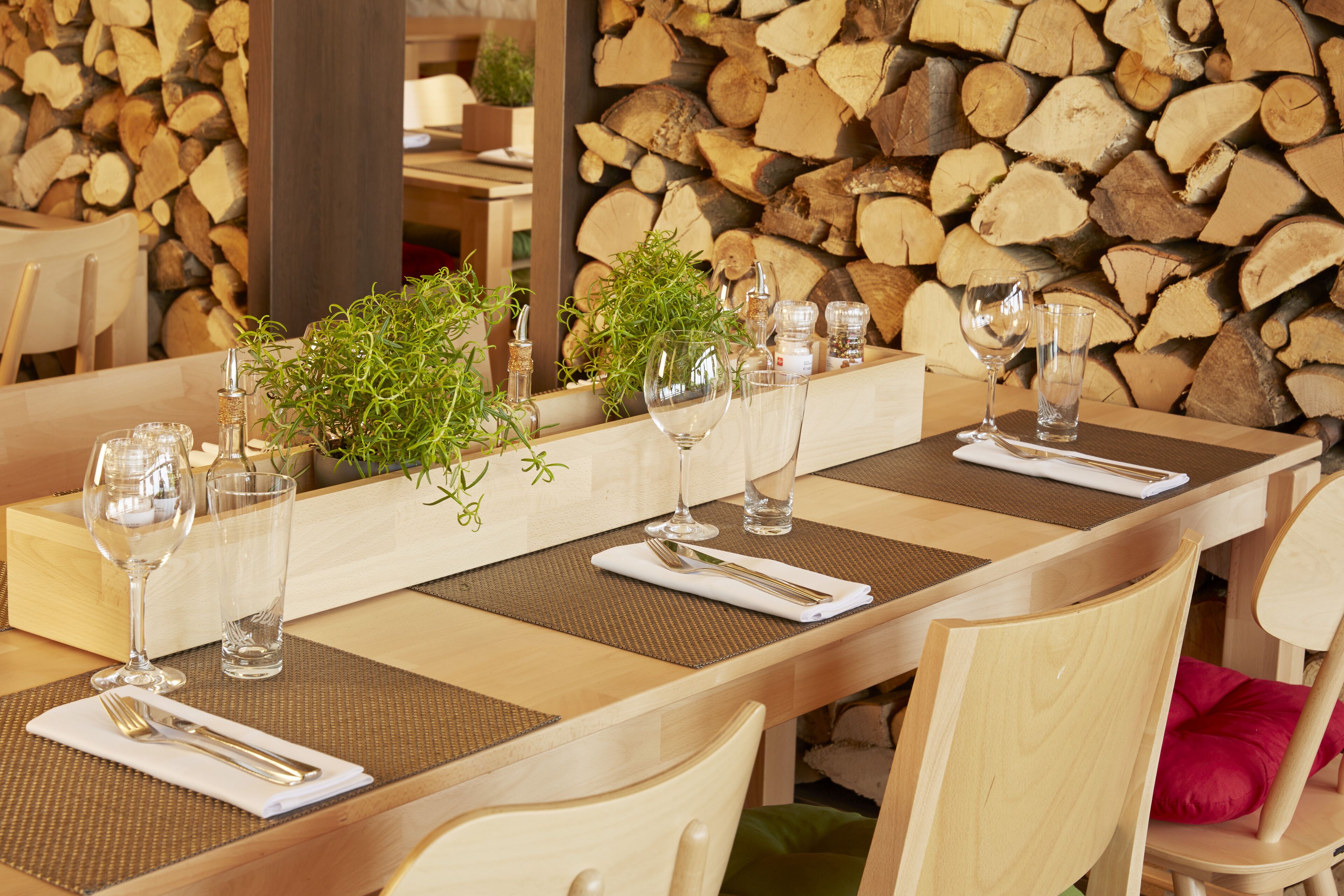 A dining table set with glassware, cutlery, and plants, with a backdrop of stacked firewood.