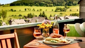 Une table avec un plateau de fromages, deux verres de vin, une bouteille et un bouquet de fleurs, le tout placé sur un balcon donnant sur un paysage verdoyant.